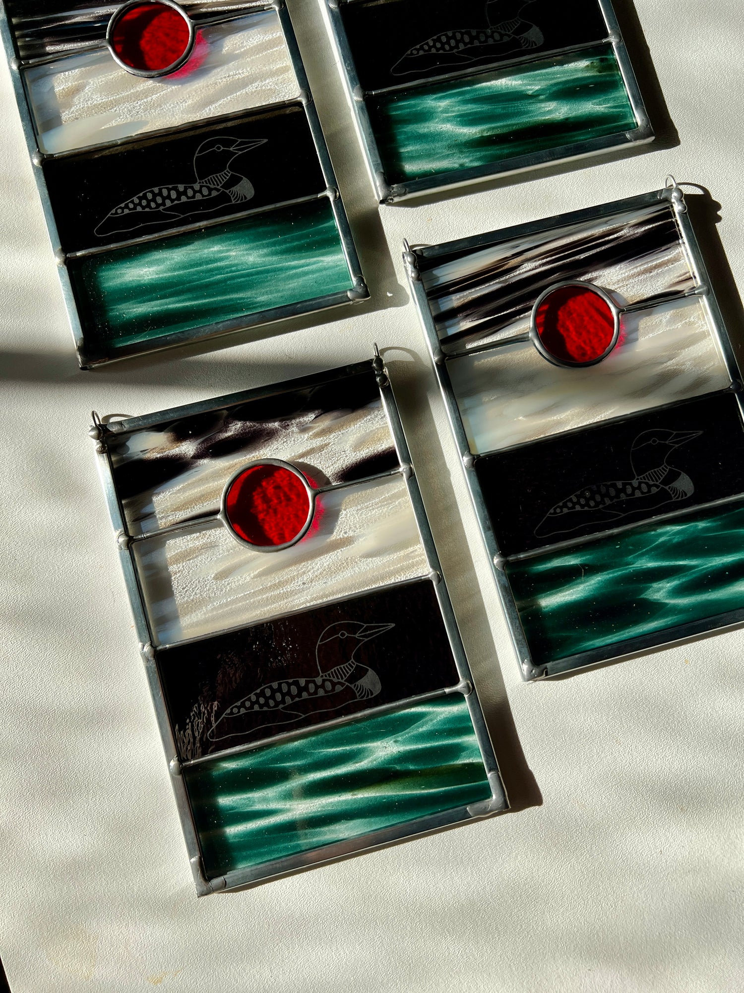 four stained glass panels with black, white, clear, teal, and red glass with loons etched on the surface, photographed at an angled on a white surface