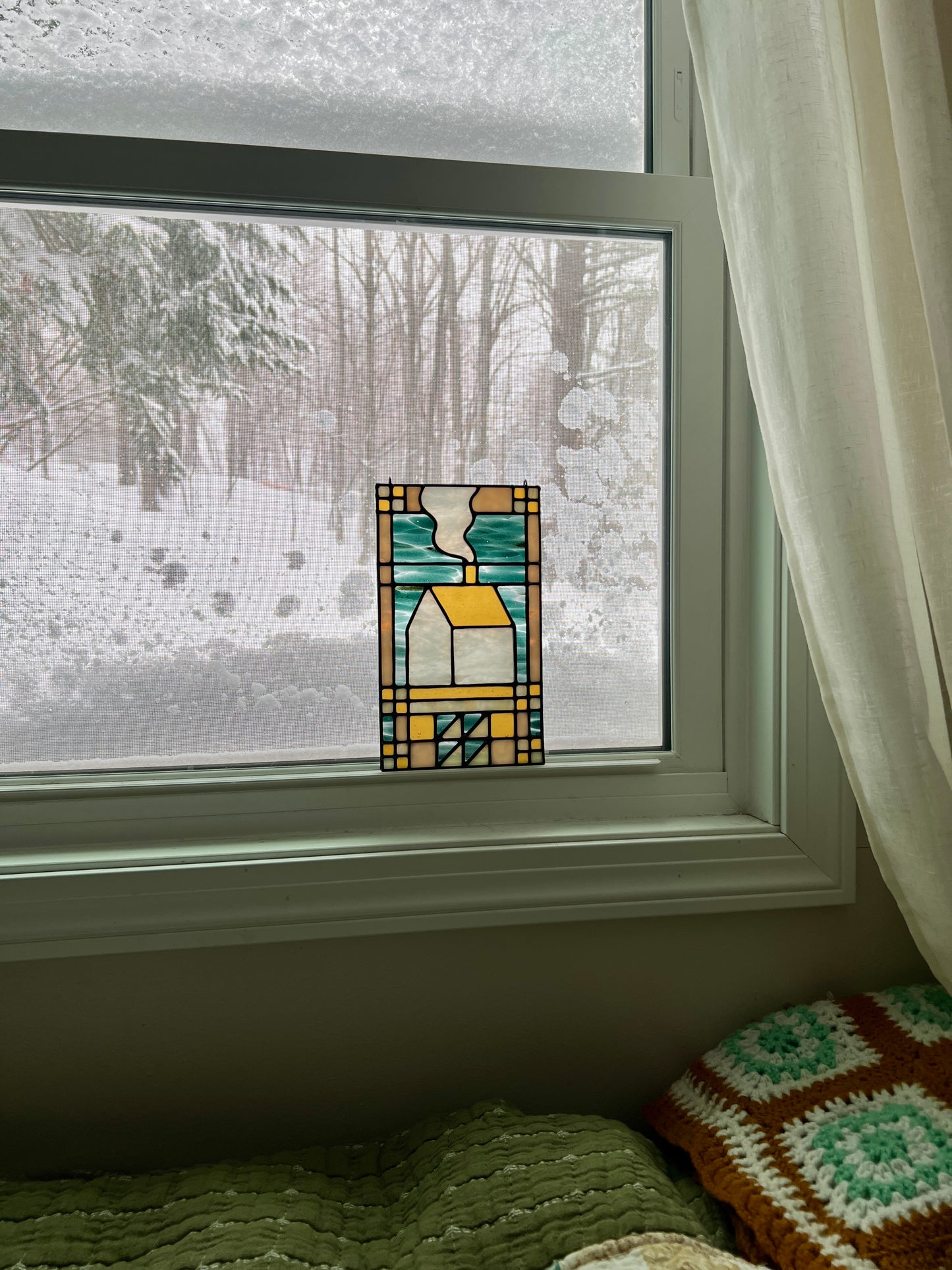 Stained glass textile inspired panel depicting a home in the center in shades of teal, amber, white, and beige, resting in a window with a snowy landscape outside and cozy blankets in the foreground