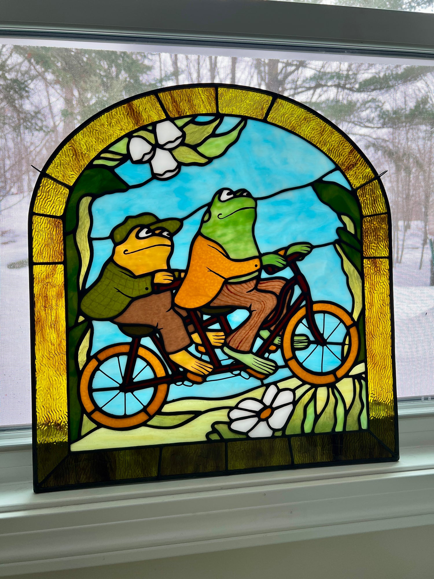 stained glass panel depicting frog and toad riding a bicycle resting in a window sill
