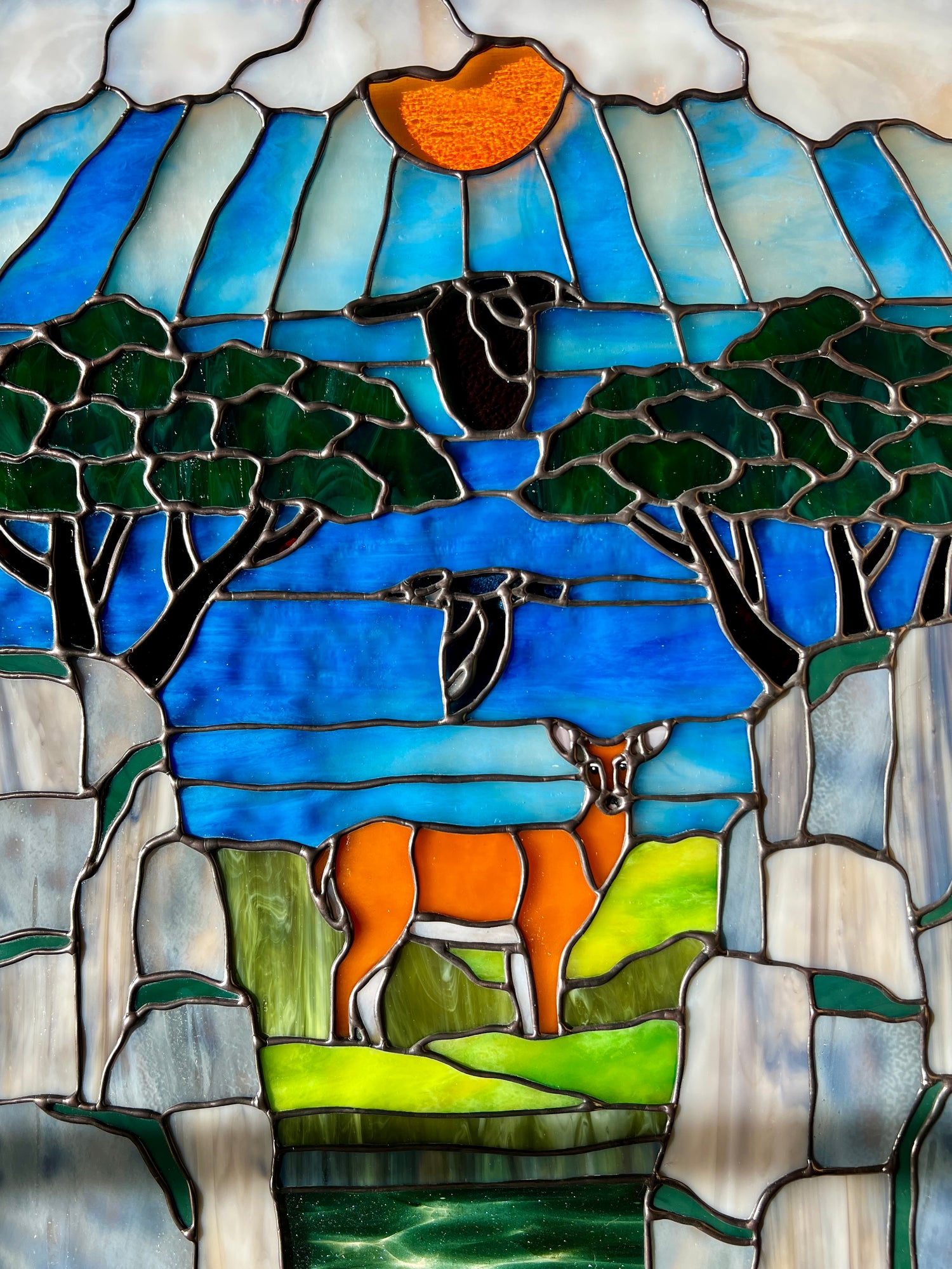large stained glass panel depicting mossy, rocky cliffs and trees, with a deer, a kingfisher, and a raven standing amongst a river, greenery, and bright blue skies with some clouds and a bright yellow orange sun.
