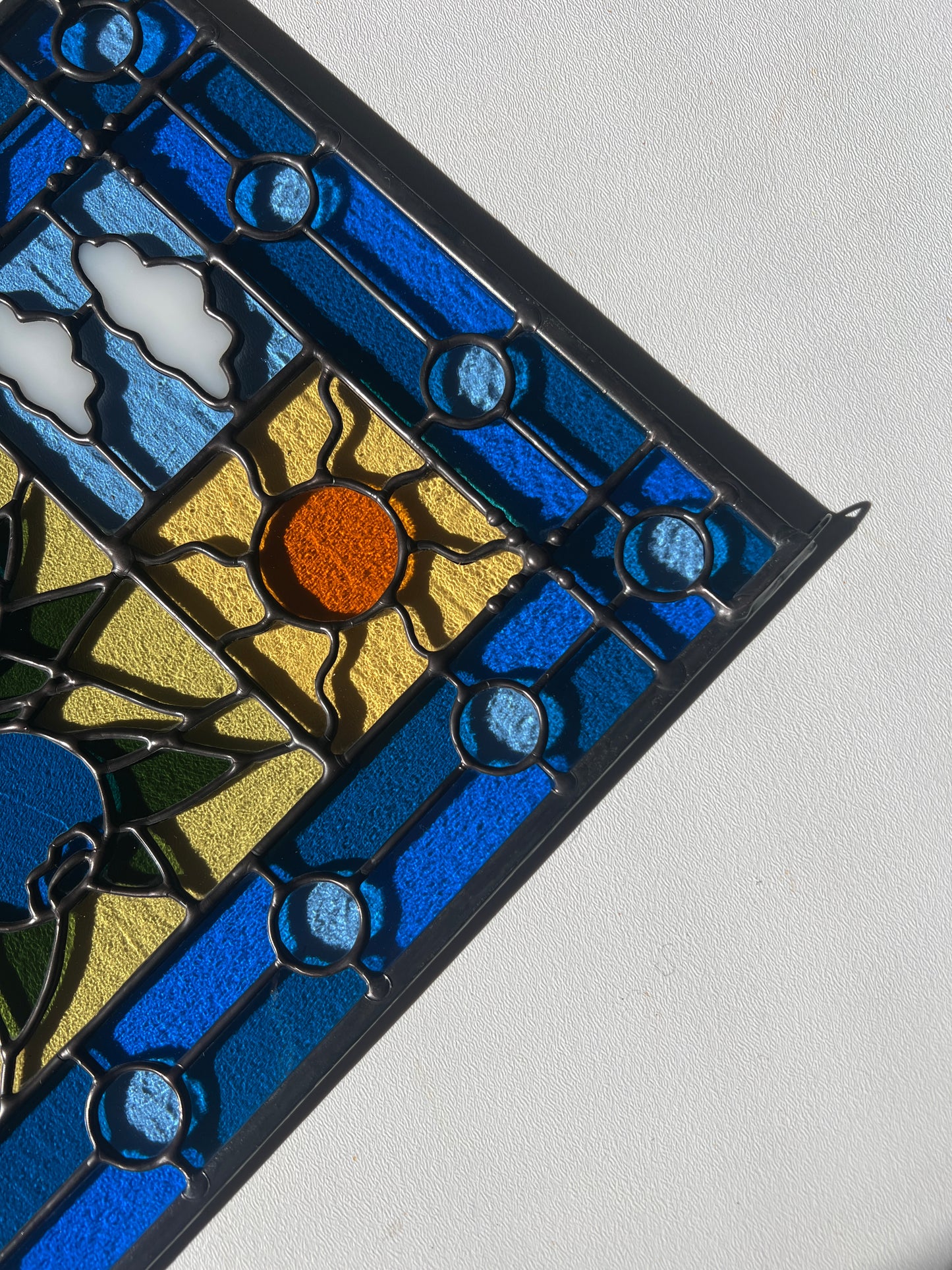 close up of a stained glass panel with a simple compartment style design featuring clouds, sun, water, and a small bunch of blueberries in the middle with a blue border