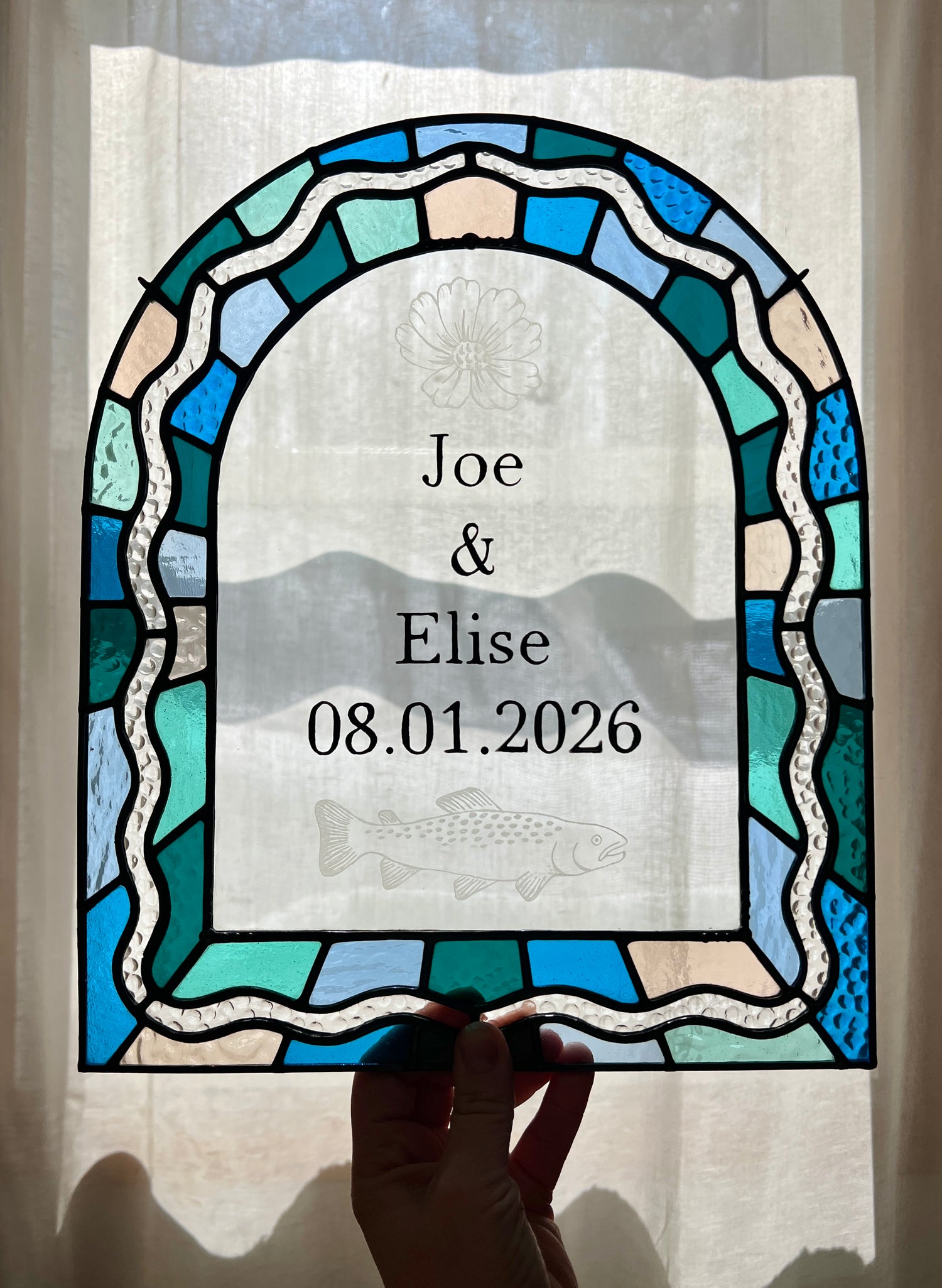 Stained glass panel in the shape of an arch with a multicolor blue, teal, turquoise, and pink border. Within the border is squiggle detail in clear textured glass. The center of the arch is clear uncolored glass, with personalized text and hand etched details of a cosmos flower and trout. The piece is held by a hand against a sheer white current, providing gentle backlighting.