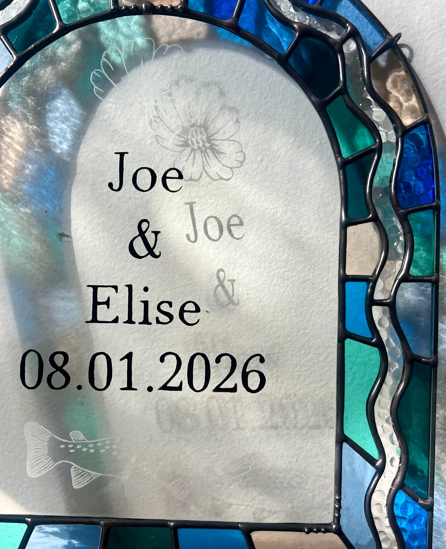 Close up of a stained glass panel in the shape of an arch with a multicolor blue, teal, turquoise, and pink border. Within the border is squiggle detail in clear textured glass. The center of the arch is clear uncolored glass, with personalized text and hand etched details of a cosmos flower and trout. The piece is held by a hand against a white wall, with gentle sunlight casting intermittent shadows and refractions.