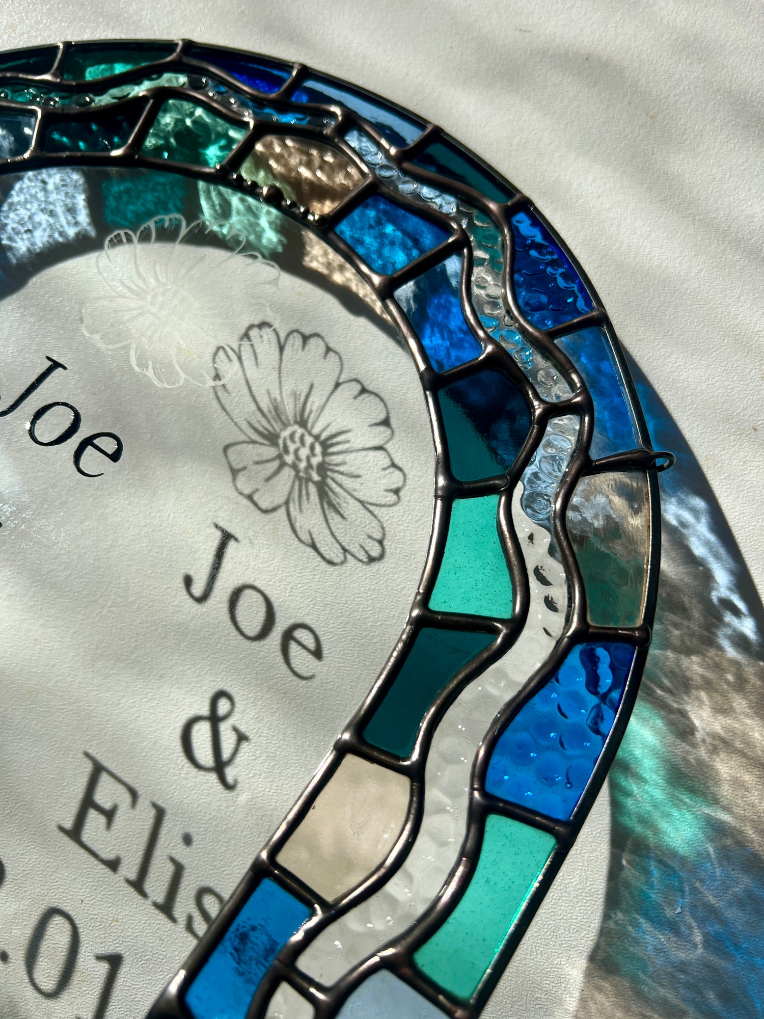 Close up of a custom stained glass piece, depicting a colorful border in shades of blue and green, and personalized hand painted texture and a hand etching of a cosmos flower and its shadow in the bright sunlight.