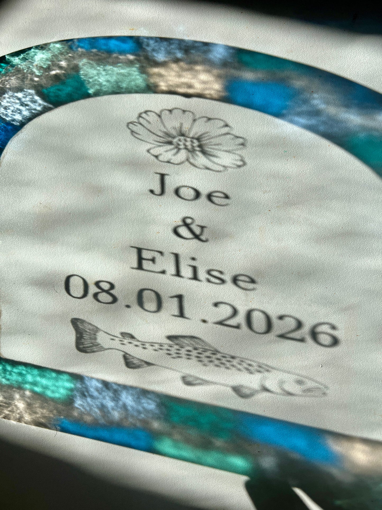 Refractions and shadows of custom text and colorful blue and teal border of a stained glass piece. The refractions are visible against a white wall. The etched illustrations are casting shadows of a cosmos flower and trout.