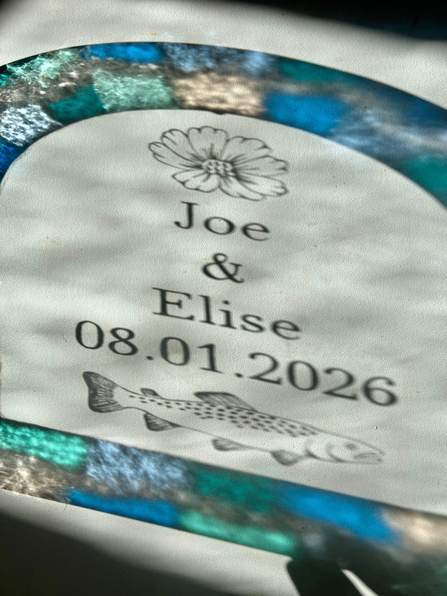 Refractions and shadows of custom text and colorful blue and teal border of a stained glass piece. The refractions are visible against a white wall. The etched illustrations are casting shadows of a cosmos flower and trout.
