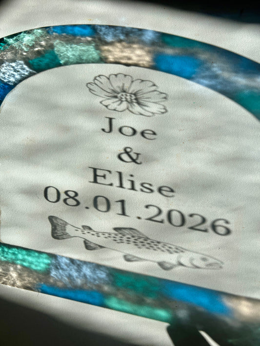 Refractions and shadows of custom text and colorful blue and teal border of a stained glass piece. The refractions are visible against a white wall. The etched illustrations are casting shadows of a cosmos flower and trout.