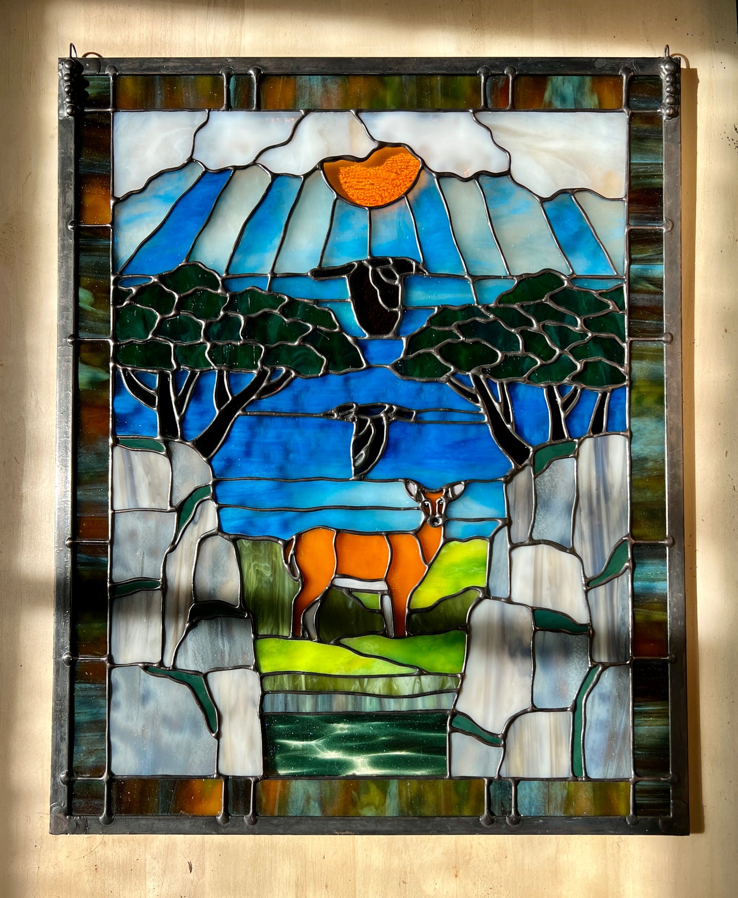 large stained glass panel depicting mossy, rocky cliffs and trees, with a deer, a kingfisher, and a raven standing amongst a river, greenery, and bright blue skies with some clouds and a bright yellow orange sun.