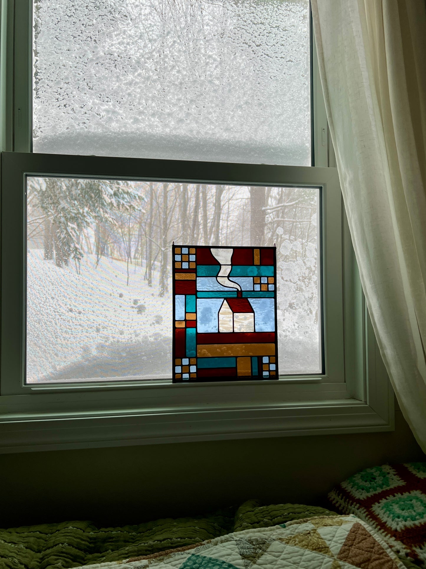 Stained glass textile inspired panel depicting a home in the center in shades of teal, brown, and blue, resting in a window with a snowy scene outside and cozy blankets in the foreground
