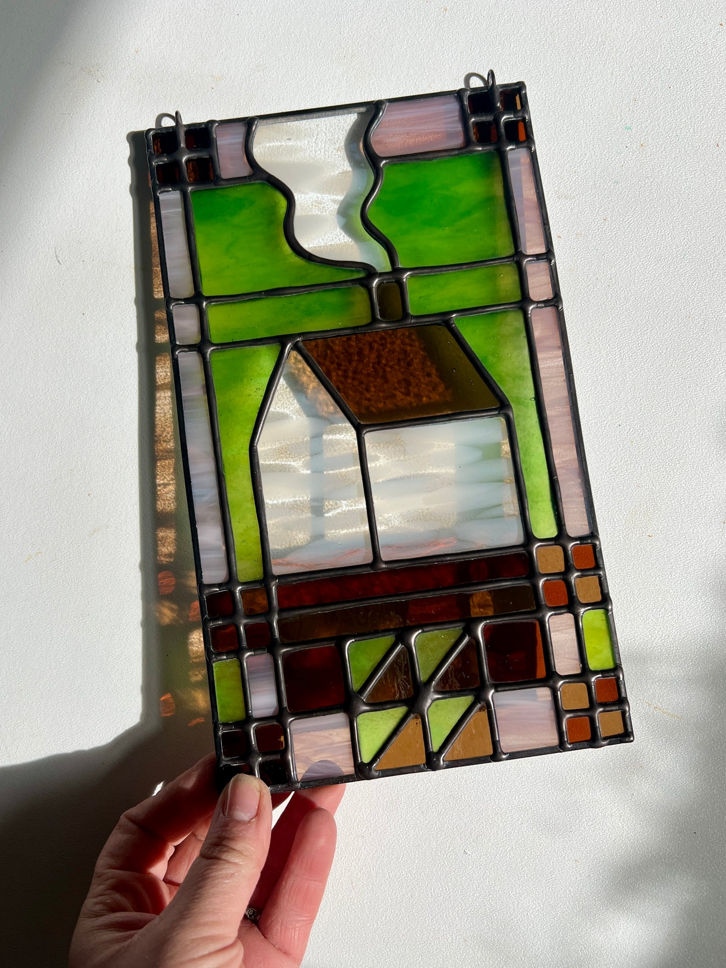 Stained glass panel with a textile inspired house design held by a hand against a white background, in shades of pink, green, brown, and white