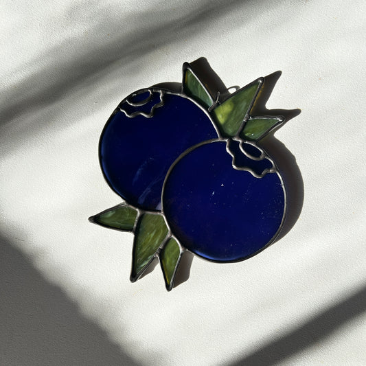a blue stained glass suncatcher depicting a small bunch of blueberries with leaves on a white surface