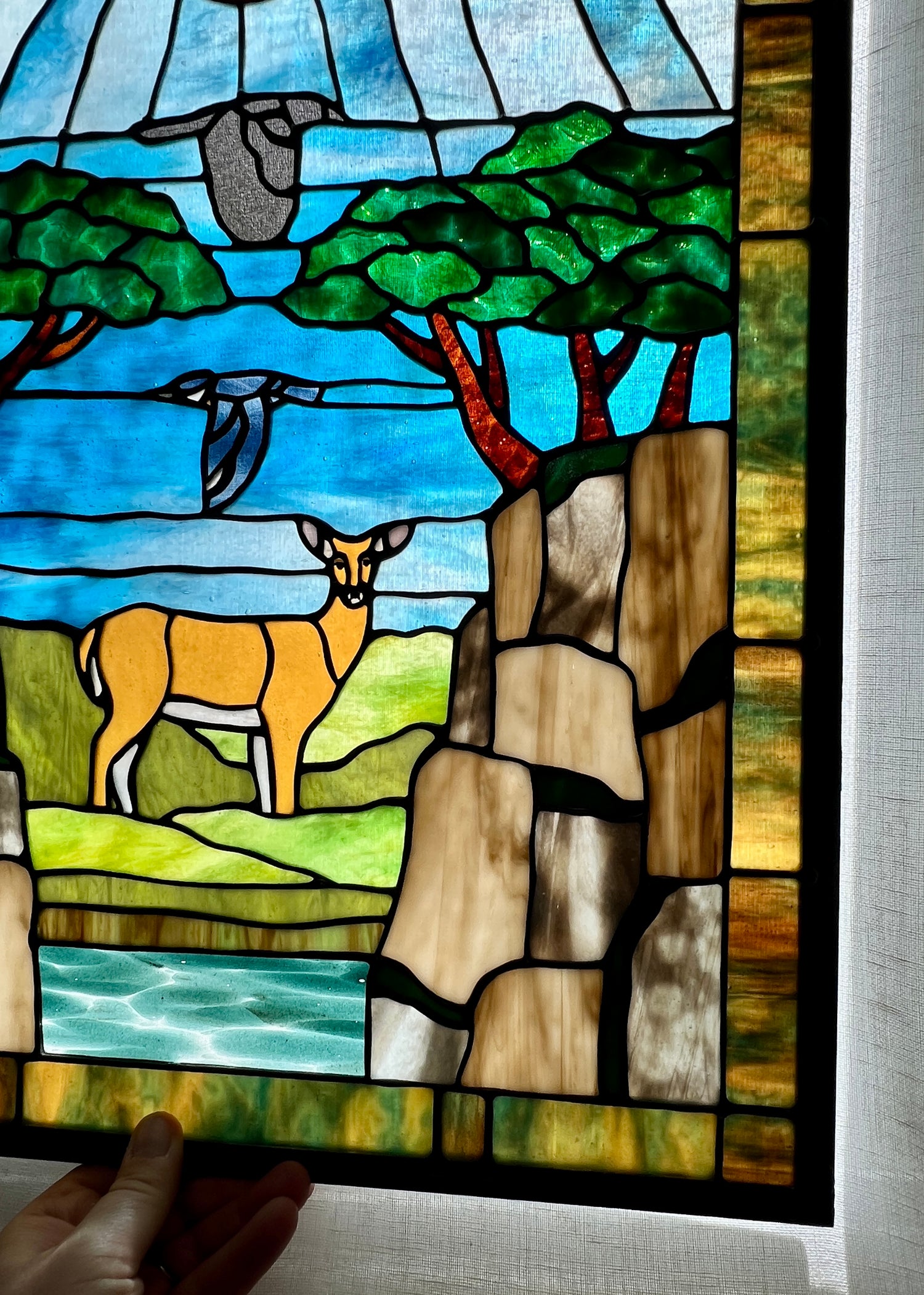 close up of a large stained glass panel depicting mossy, rocky cliffs and trees, with a deer, a kingfisher, and a raven standing amongst a river, greenery, and bright blue skies with some clouds and a bright yellow orange sun.