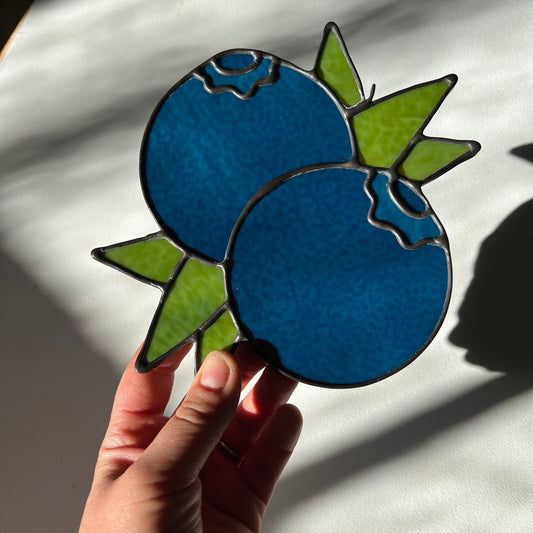 hand holding a dark blue stained glass suncatcher depicting a small bunch of blueberries with leaves
