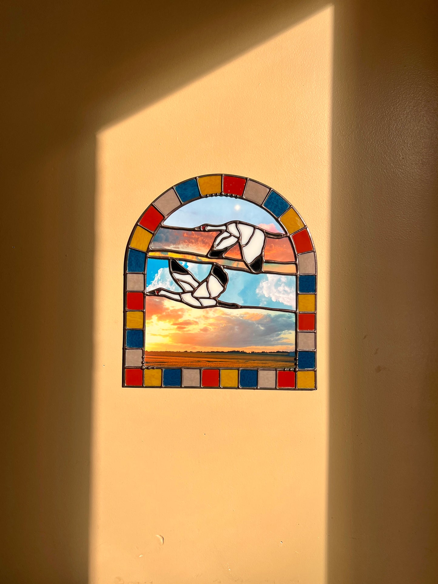 Stained glass panel with a landscape design created by collaged photographs and two cranes flying against a beige wall with bright sunlight highlighting the panel