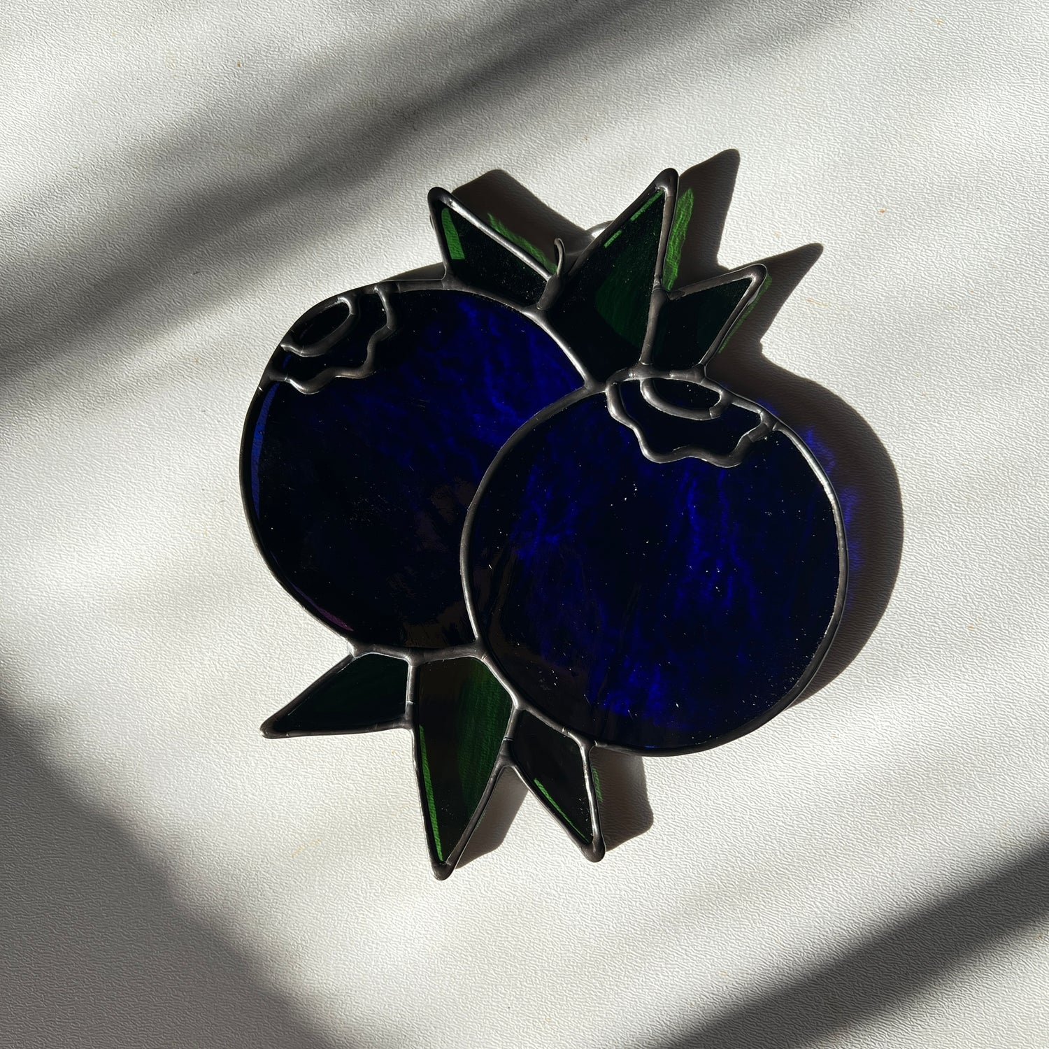 a wispy dark blue stained glass suncatcher depicting a small bunch of blueberries with leaves resting on white surface