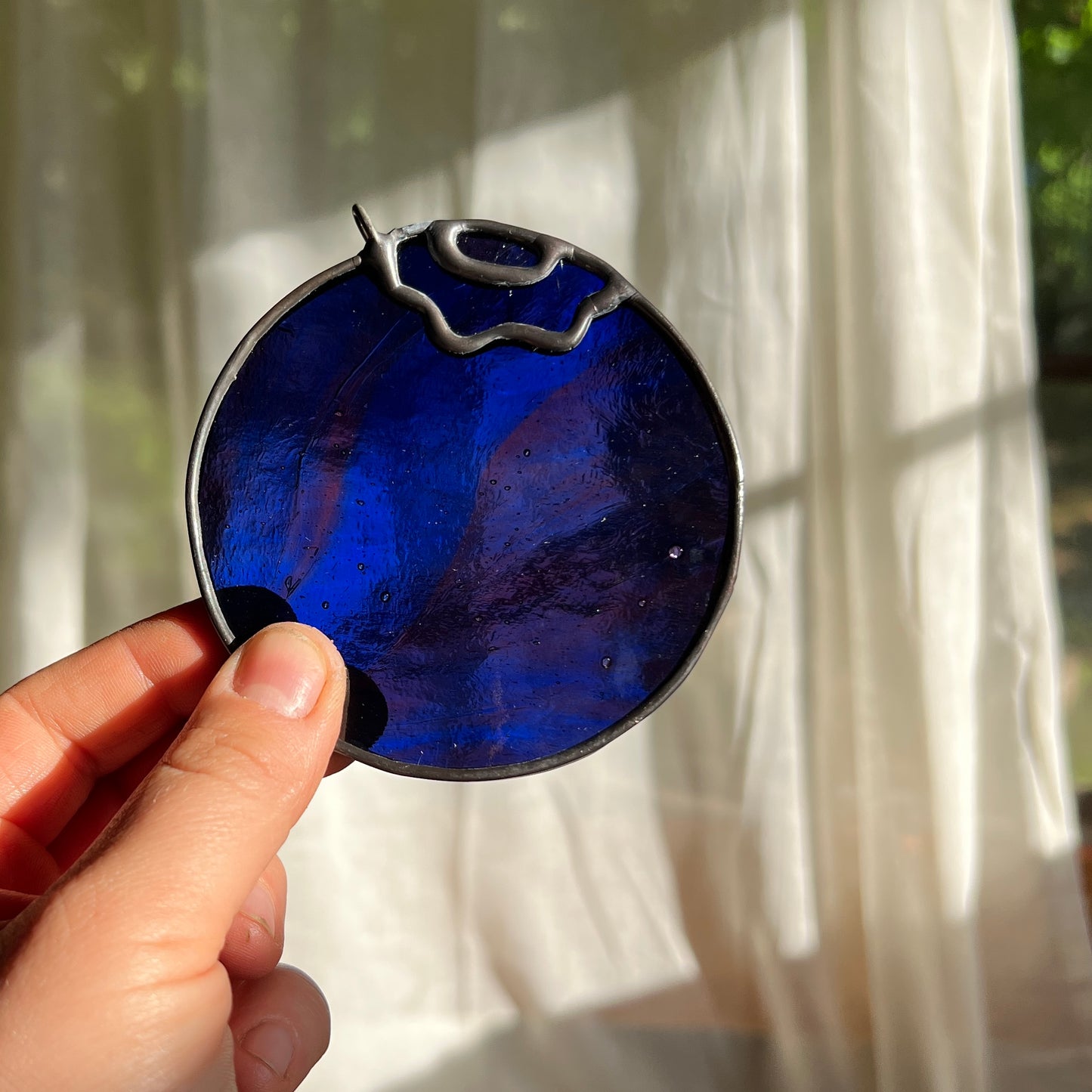 hand holding a wispy deep blue and magenta stained glass blueberry in front of a window