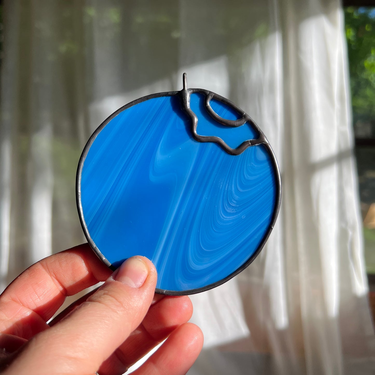 a hand holding a small streaky blue blueberry suncatcher in front of a window