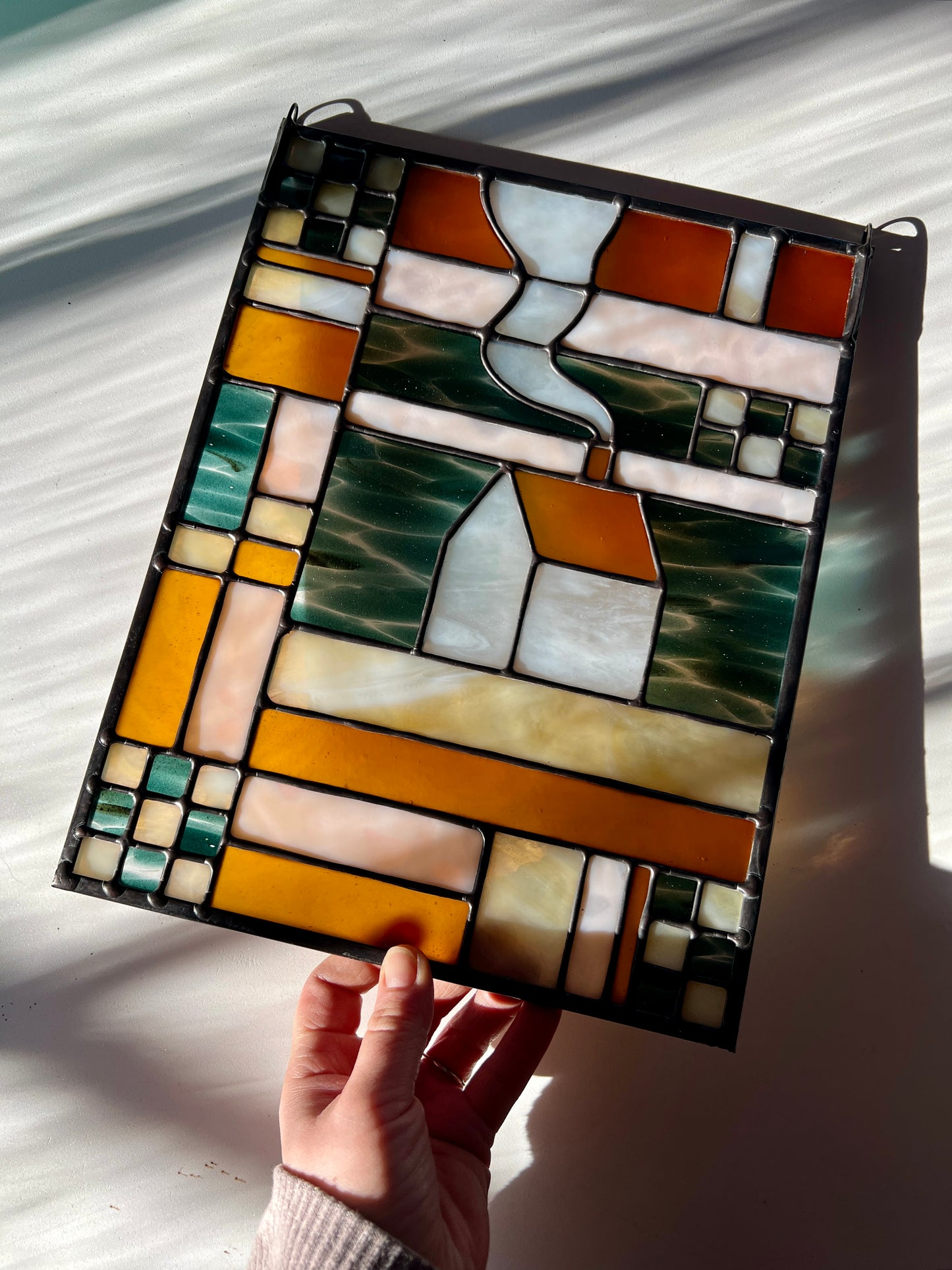 Stained glass textile inspired panel depicting a home in the center in shades of teal, amber, white, and beige, held by a hand against a white surface with sunlight and shadows cast across