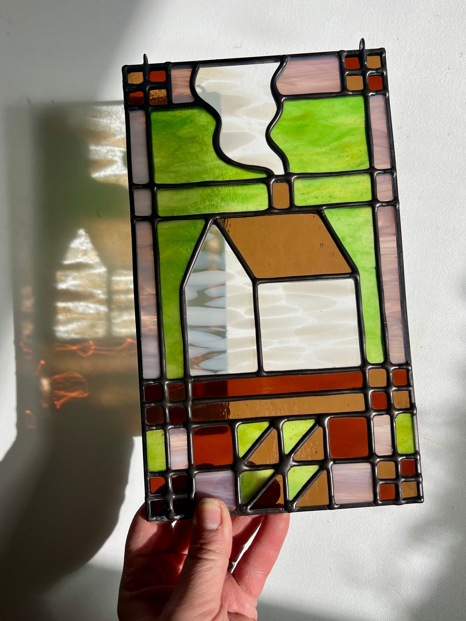 Stained glass panel with a textile inspired house design held by a hand against a white background, with bright sunlight casting refractions