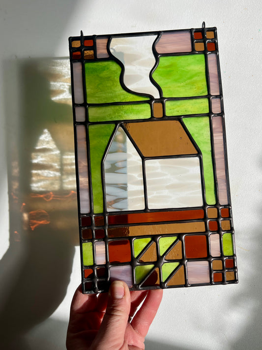 Stained glass panel with a textile inspired house design held by a hand against a white background, with bright sunlight casting refractions