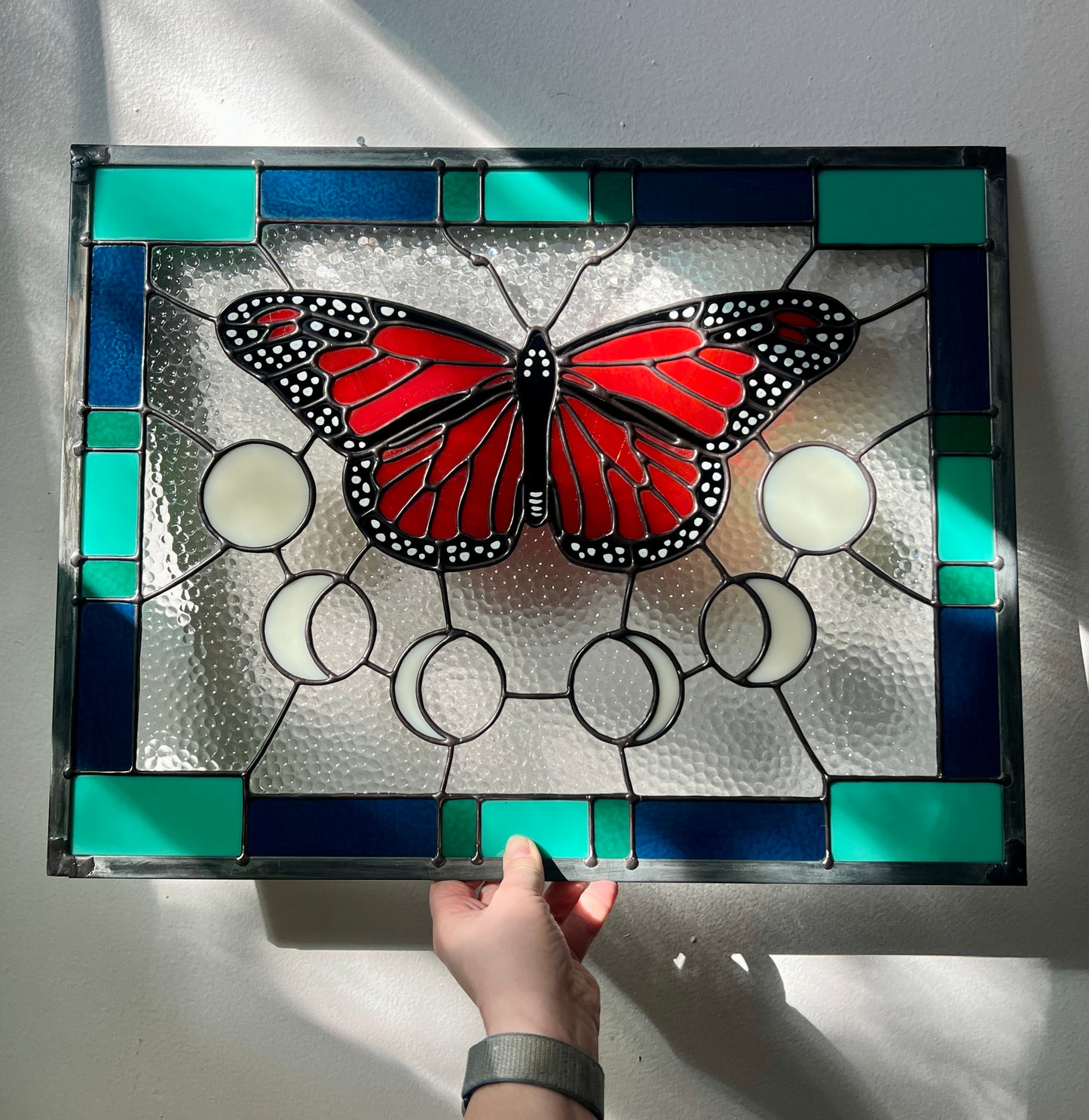 A rectangular stained glass panel with a thick blue and green geometric border and a monarch butterfly and moon phases in the center, with textured clear glass in the background. The piece is held by a hand and resting against a white wall, with soft sunlight flowing across it.