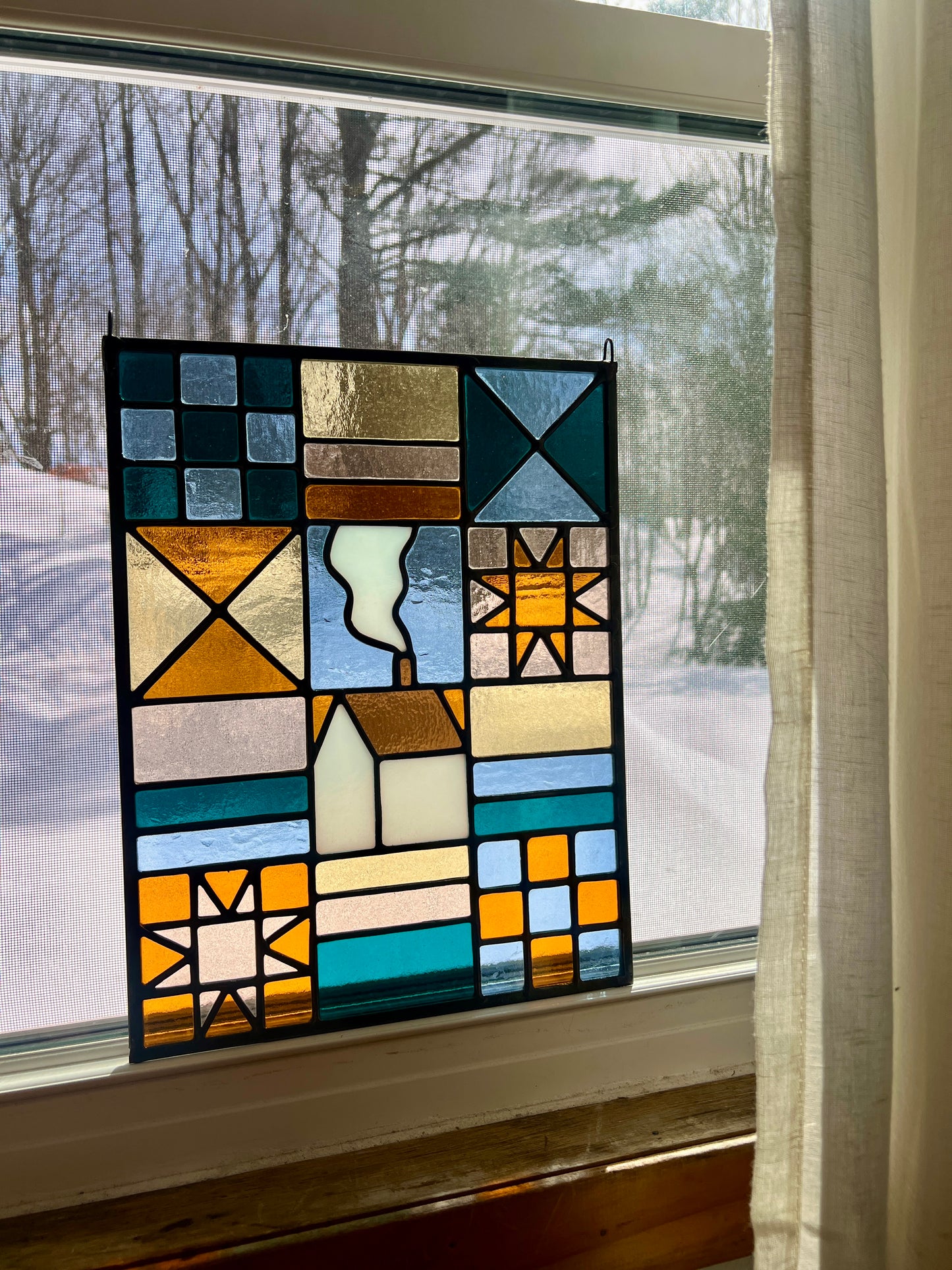 Stained glass textile inspired panel depicting a home in the center in shades of teal, pink, white, blue, and yellow, resting in a sunlit window with a snowy landscape outside