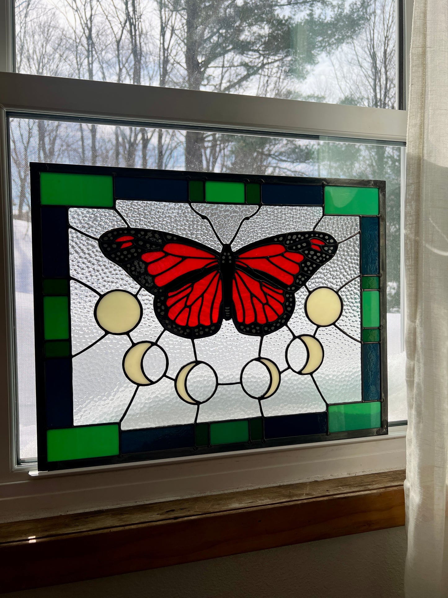 A rectangular stained glass panel with a thick blue and green geometric border and a monarch butterfly and moon phases in the center, with textured clear glass in the background. The panel is resting in a window with backlighting.