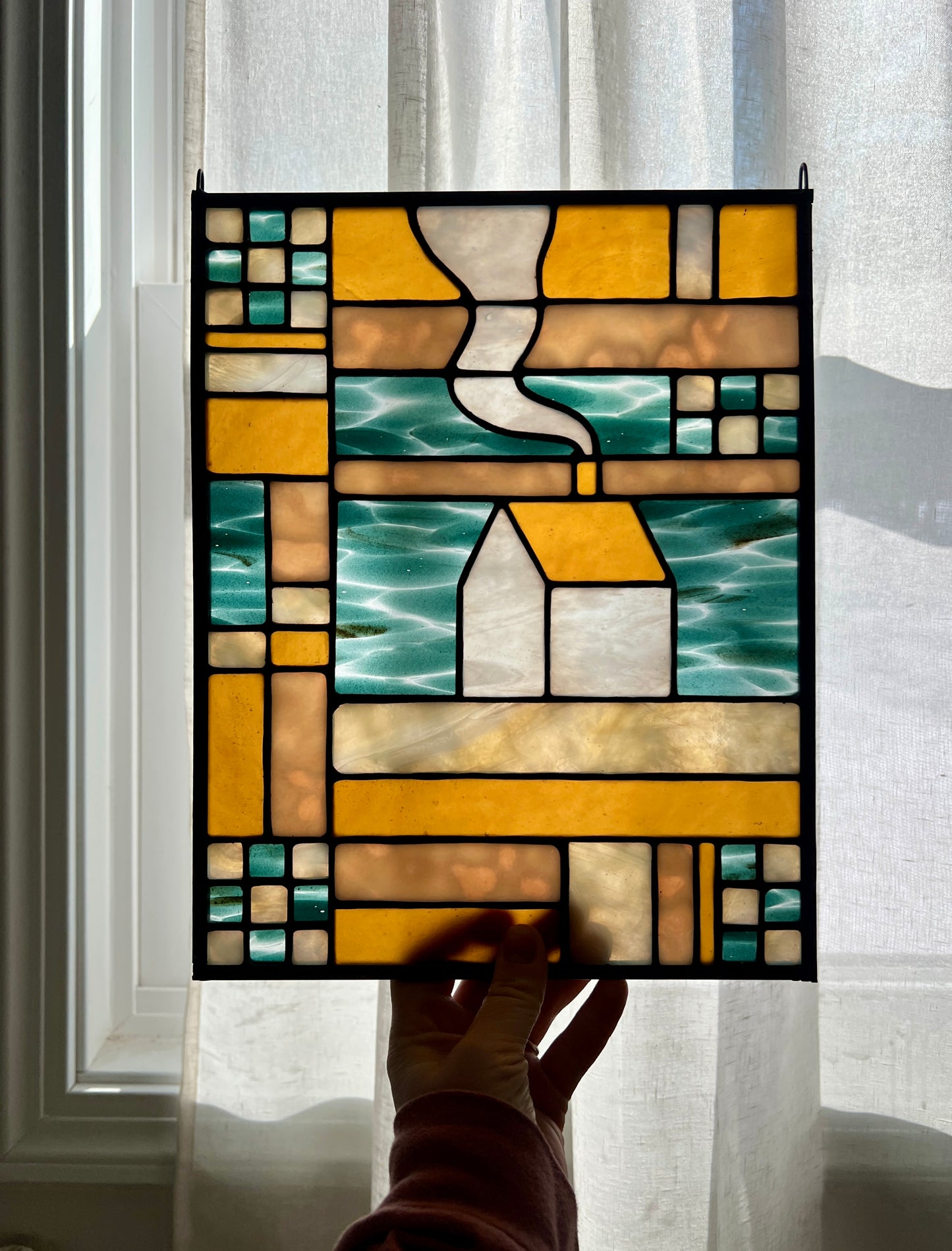 Stained glass textile inspired panel depicting a home in the center in shades of teal, amber, white, and beige, held by a hand against a brightly backlit sheer white curtain