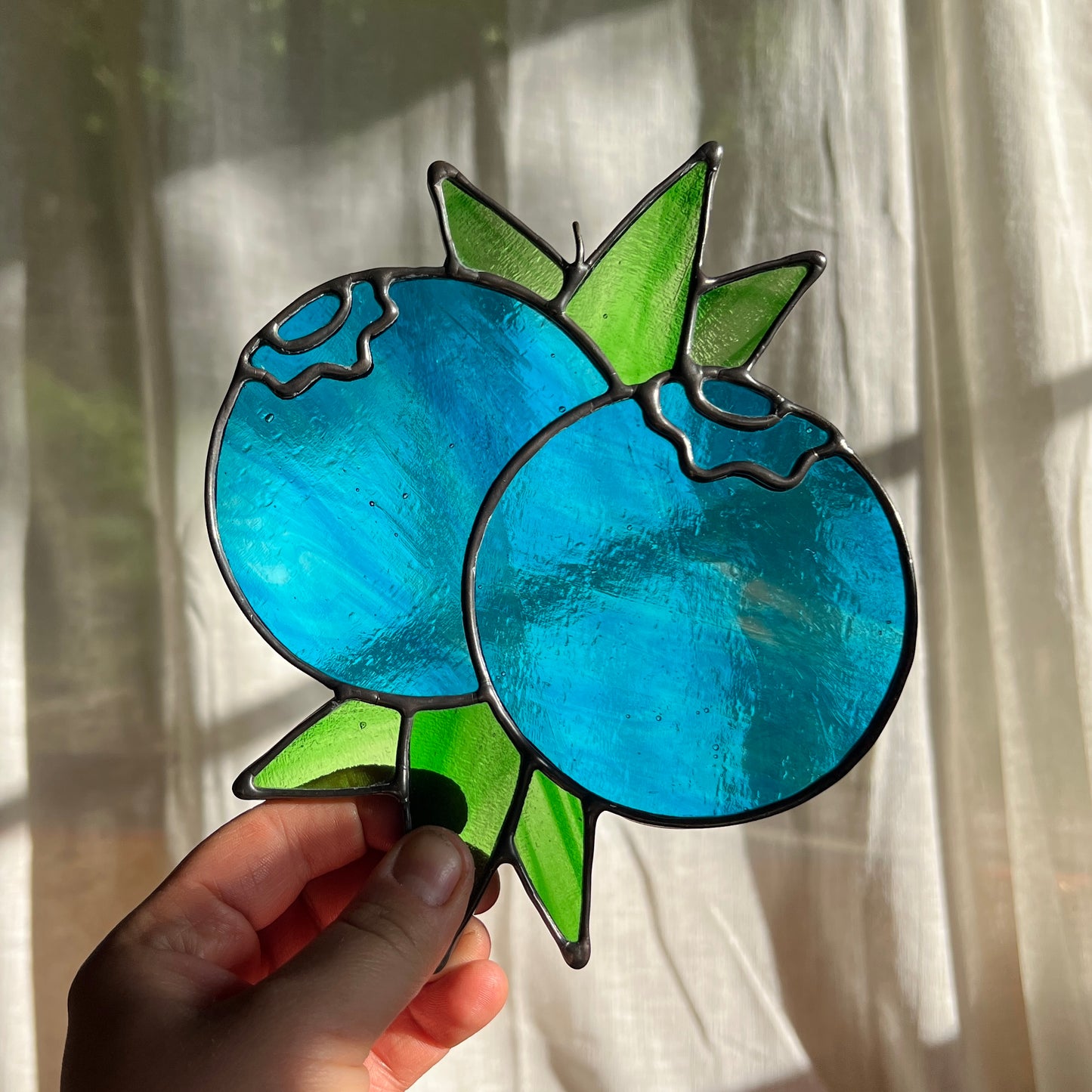 a hand holding a wispy blue suncatcher depicting a small bunch of blueberries with leaves in front of a window