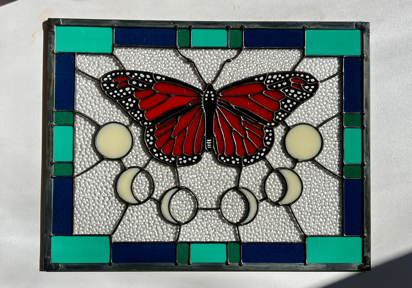 A large stained glass panel with a thick blue and green geometric border and a monarch butterfly and moon phases in the center, with textured clear glass in the background. Photographed against a white background with bright sunlight flowing across it.