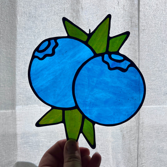 A hand holding a stained glass suncatcher depicting two blueberries and some leaves against a gently backlit white background.