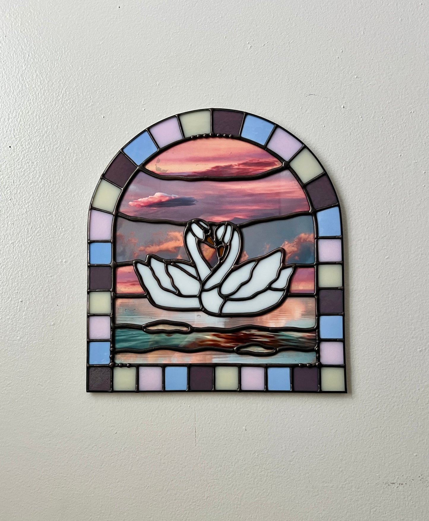 Stained glass panel with a colorful pastel arch border and two white swans in the center. Collaged photographs are used to create a sunset background. The piece is hanging up against a white wall.