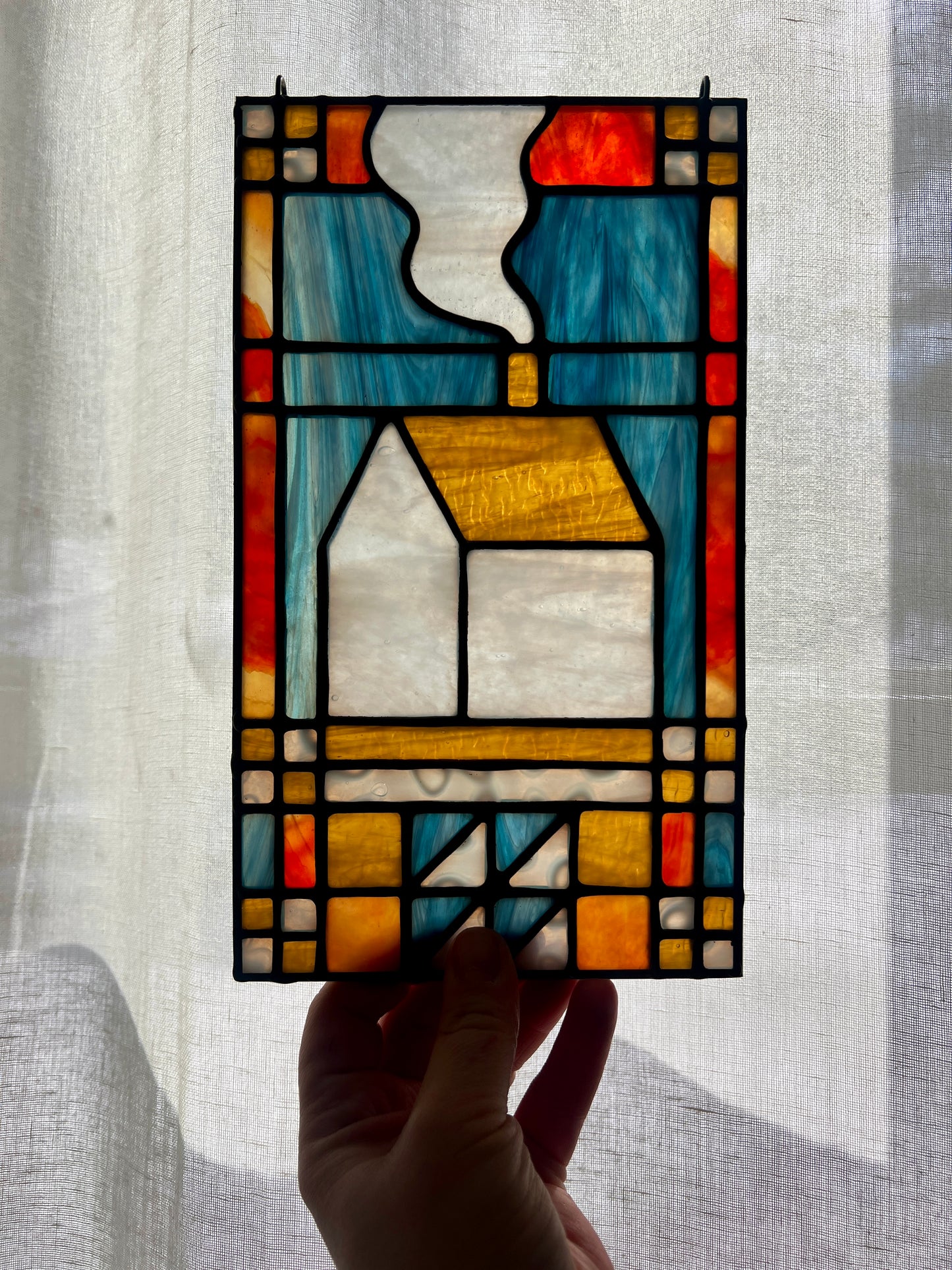 Stained glass textile inspired panel depicting a home in the center in shades of red, blue, white, and amber, held by a hand in front of a sheer white brightly backlit curtain