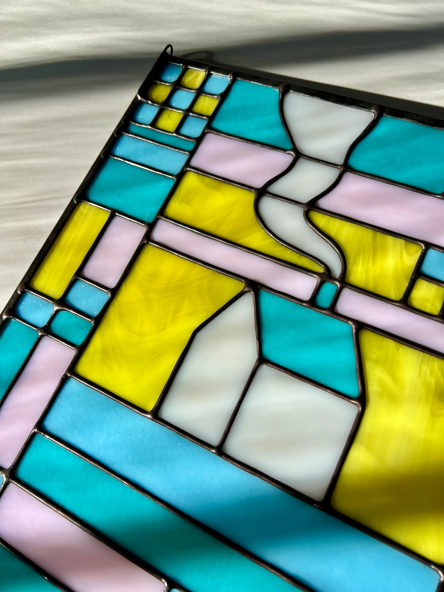 Close up of a stained glass textile inspired panel depicting a home in the center in shades of teal, pink, white, blue, and yellow,