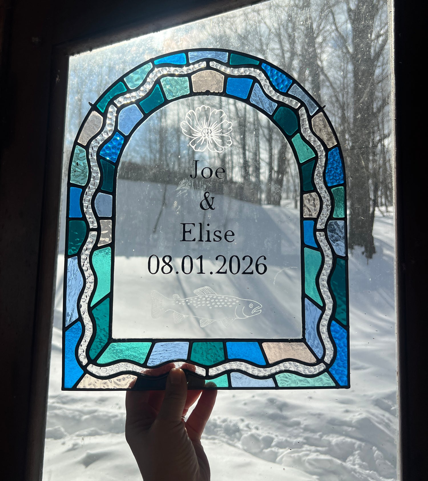 Stained glass panel in the shape of an arch with a multicolor blue, teal, turquoise, and pink border. Within the border is squiggle detail in clear textured glass. The center of the arch is clear uncolored glass, with personalized text and hand etched details of a cosmos flower and trout. The piece is held by a hand up to a window with a winter scene outside.