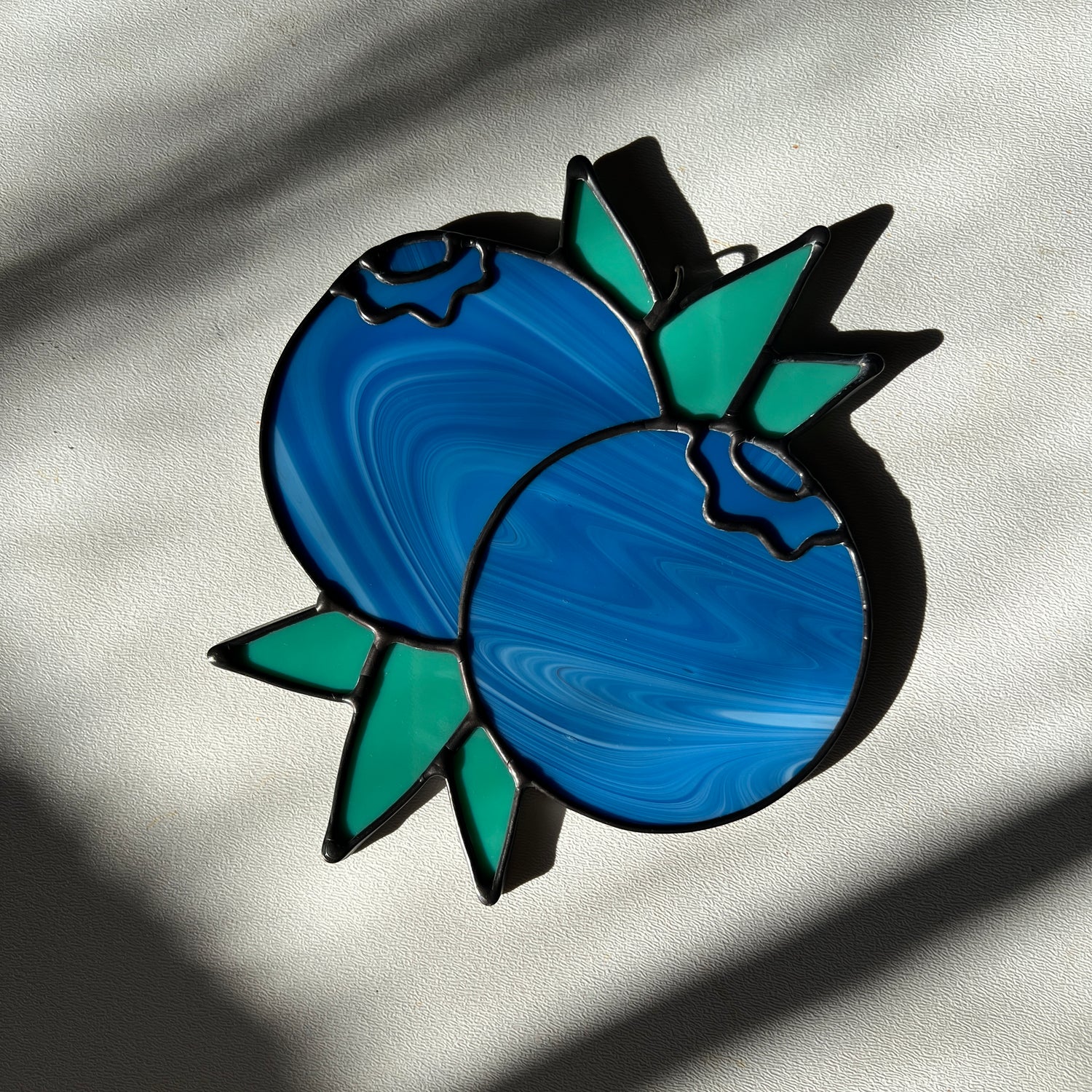 a streaky blue stained glass suncatcher depicting a small bunch of blueberries with leaves resting on a white surface