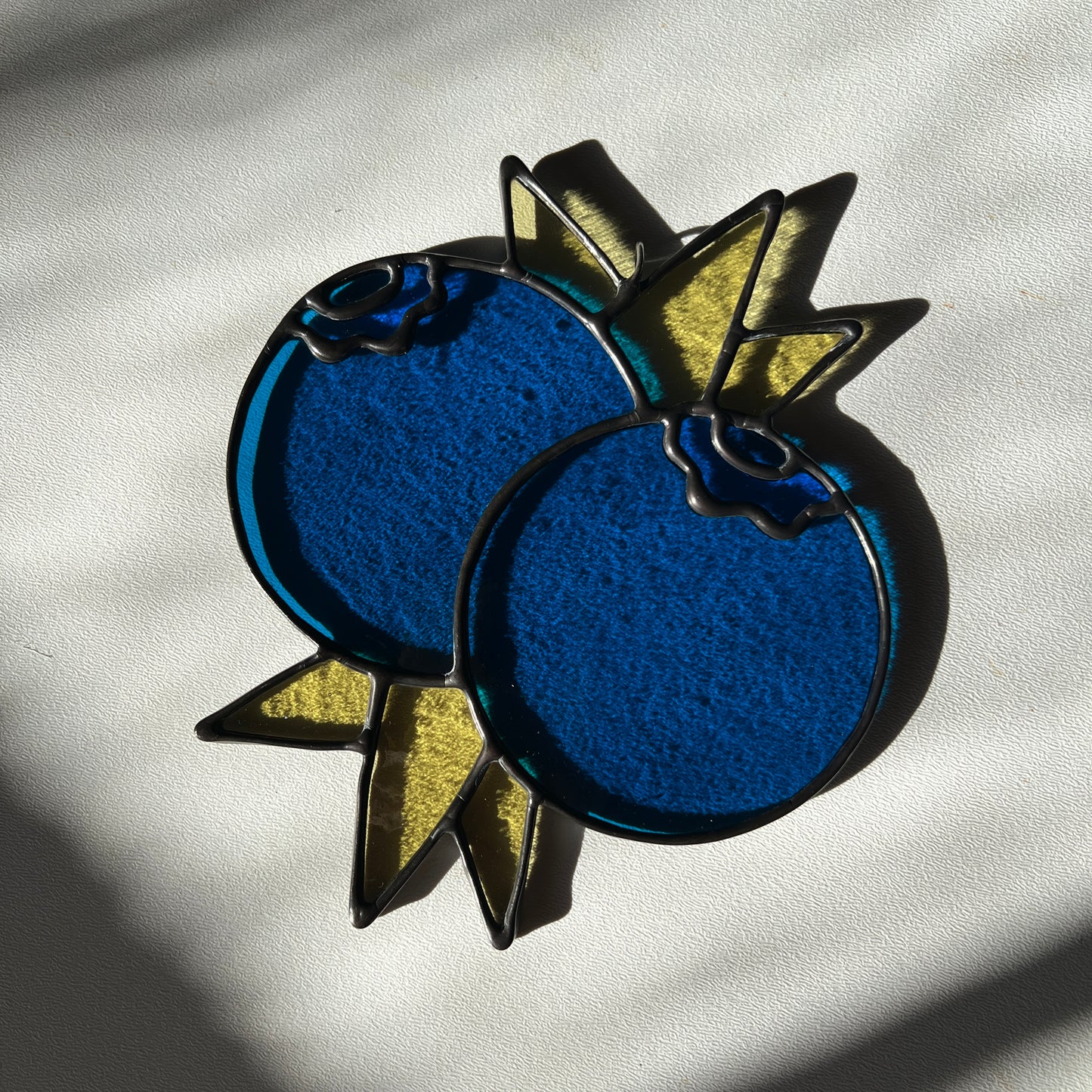 a stained glass suncatcher depicting a small bunch of blueberries with leaves resting on a white surface
