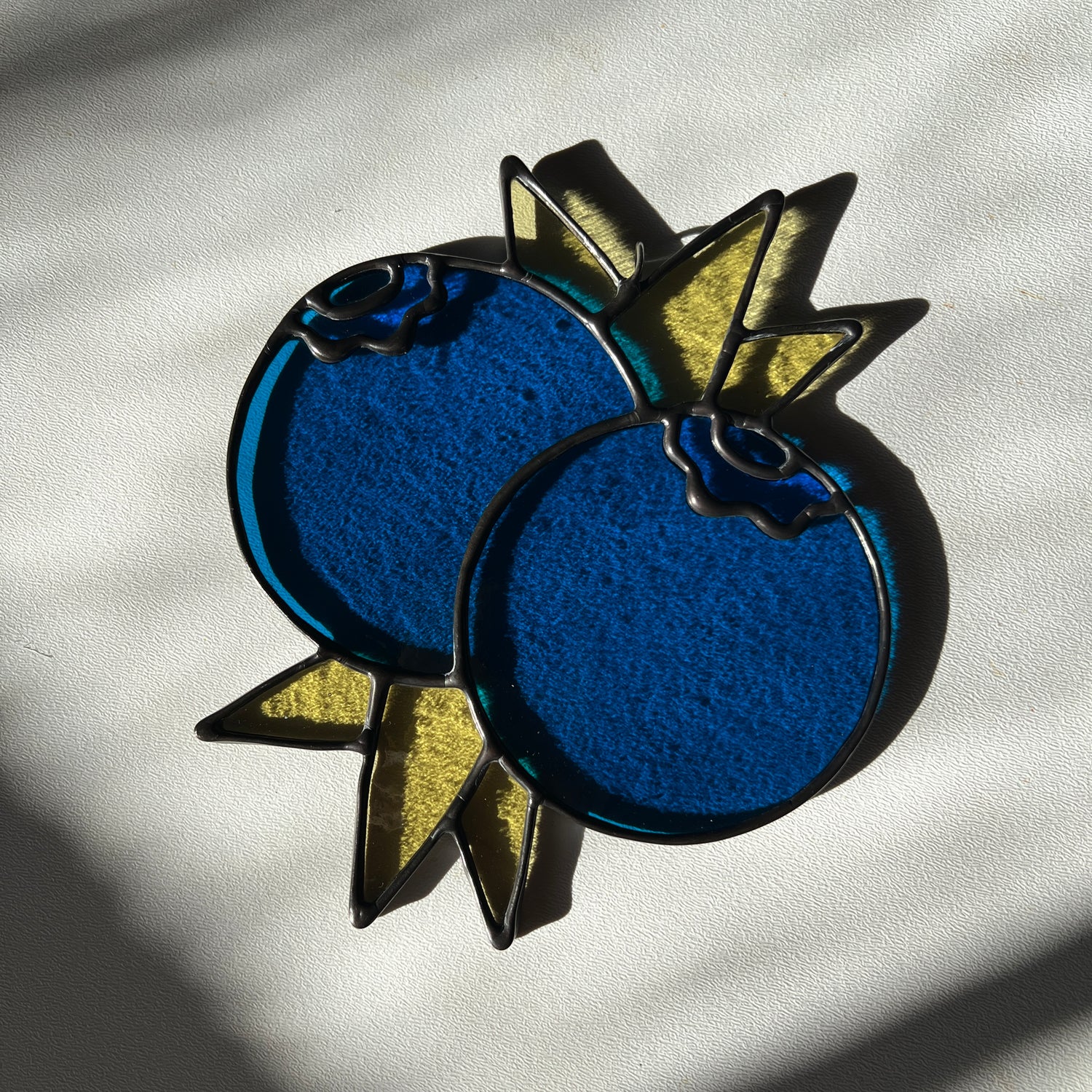 a stained glass suncatcher depicting a small bunch of blueberries with leaves resting on a white surface