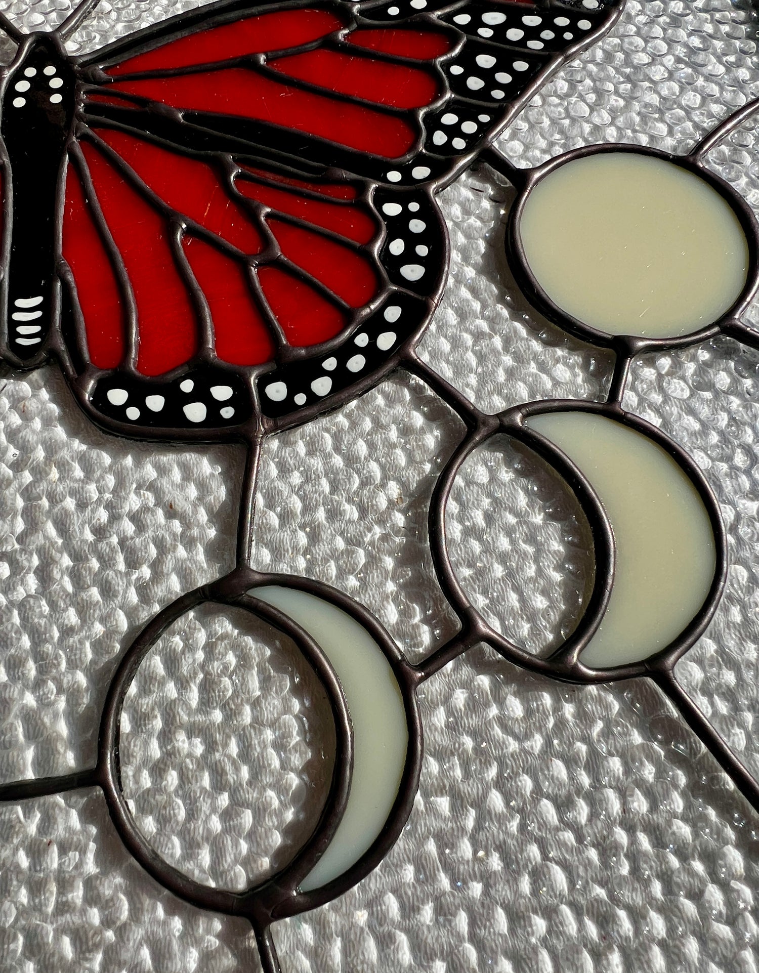 Close up of a stained glass panel depicting hand painted details of the white accents of a monarch butterfly and the smooth solder of moons arranged in a moon phase progression, with textured clear glass as the background.
