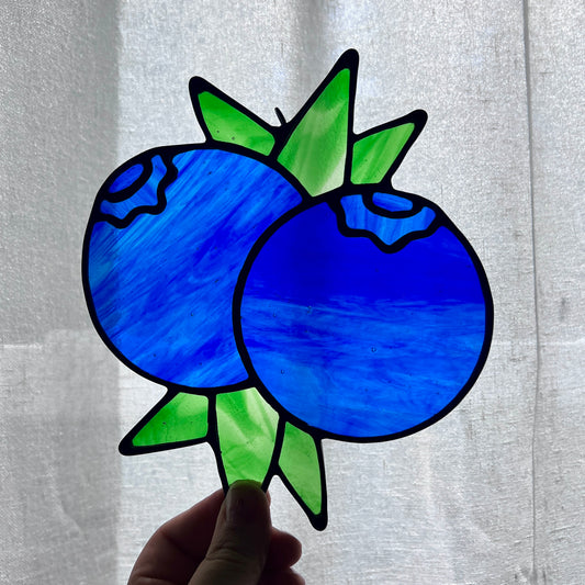 A hand holding a stained glass suncatcher depicting two blueberries and some leaves against a gently backlit white background.