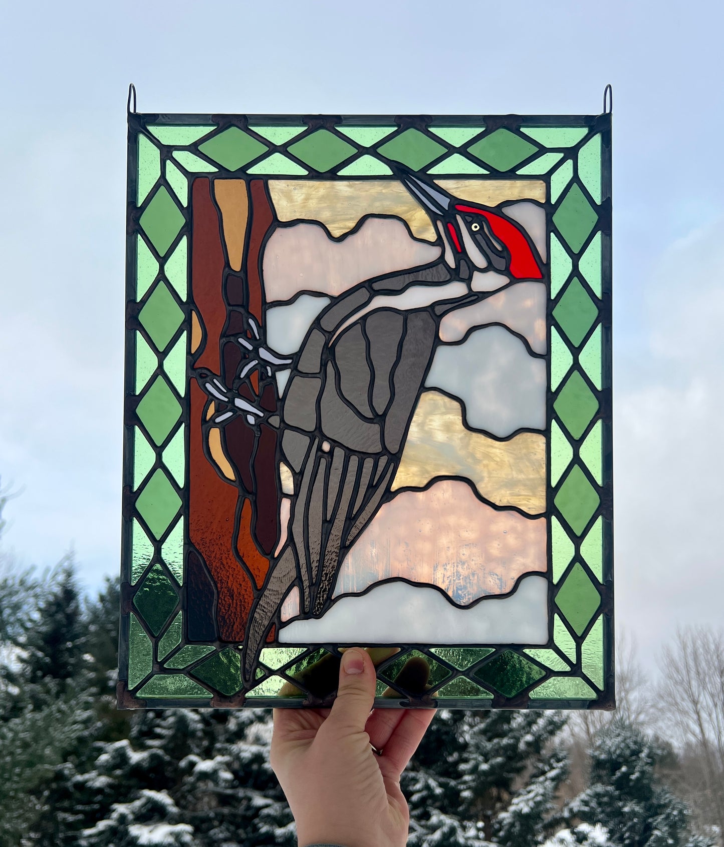 stained glass panel depicting a pileated woodpecker with a green diamond pattern border, held up in someone's hand against a wintery backdrop