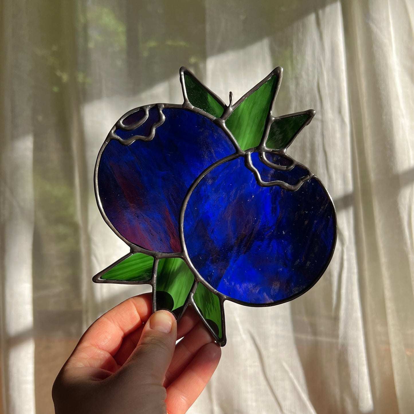 hand holding a wispy blue stained glass suncatcher depicting a small bunch of blueberries with leaves in front of a window