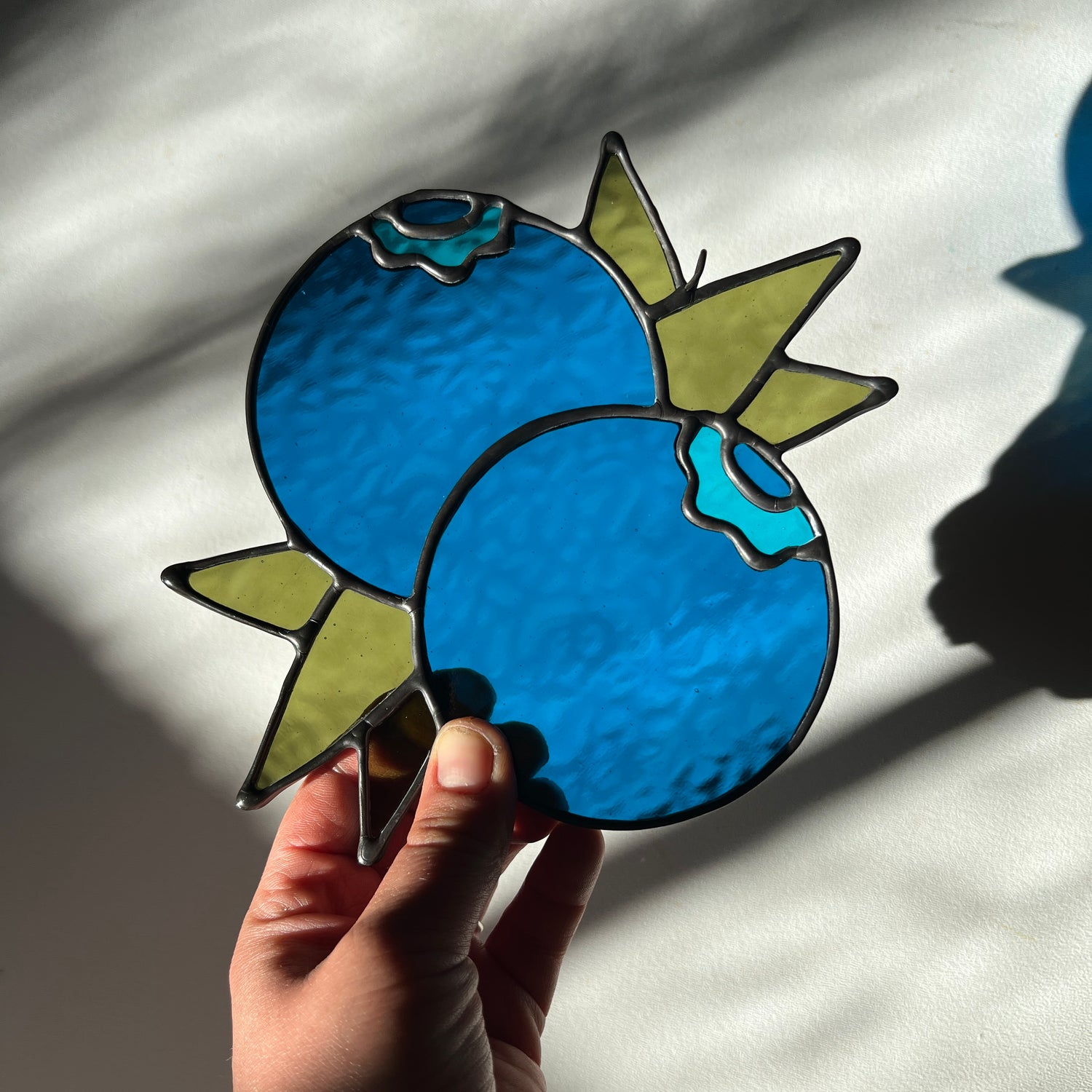 hand holding a textured blue stained glass suncatcher depicting a small bunch of blueberries with leaves
