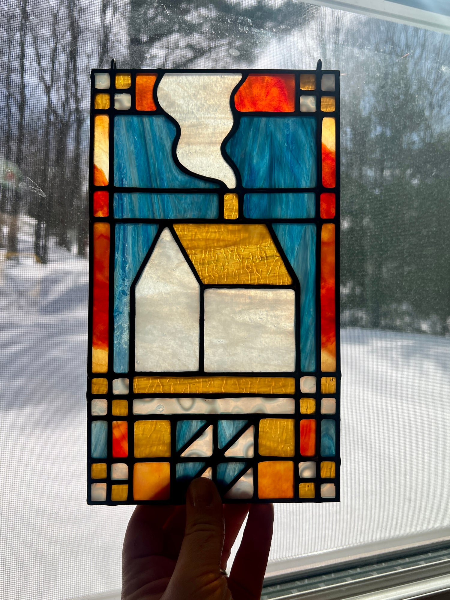 Stained glass textile inspired panel depicting a home in the center in shades of red, blue, white, and amber, held by a hand in front of a window with a snowy landscape outside