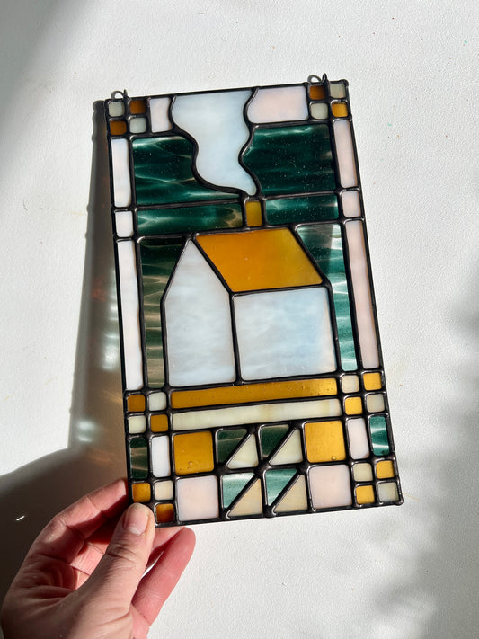 Stained glass textile inspired panel depicting a home in the center in shades of teal, amber, white, and beige, held by a hand against a white surface with bright sunlight cast across