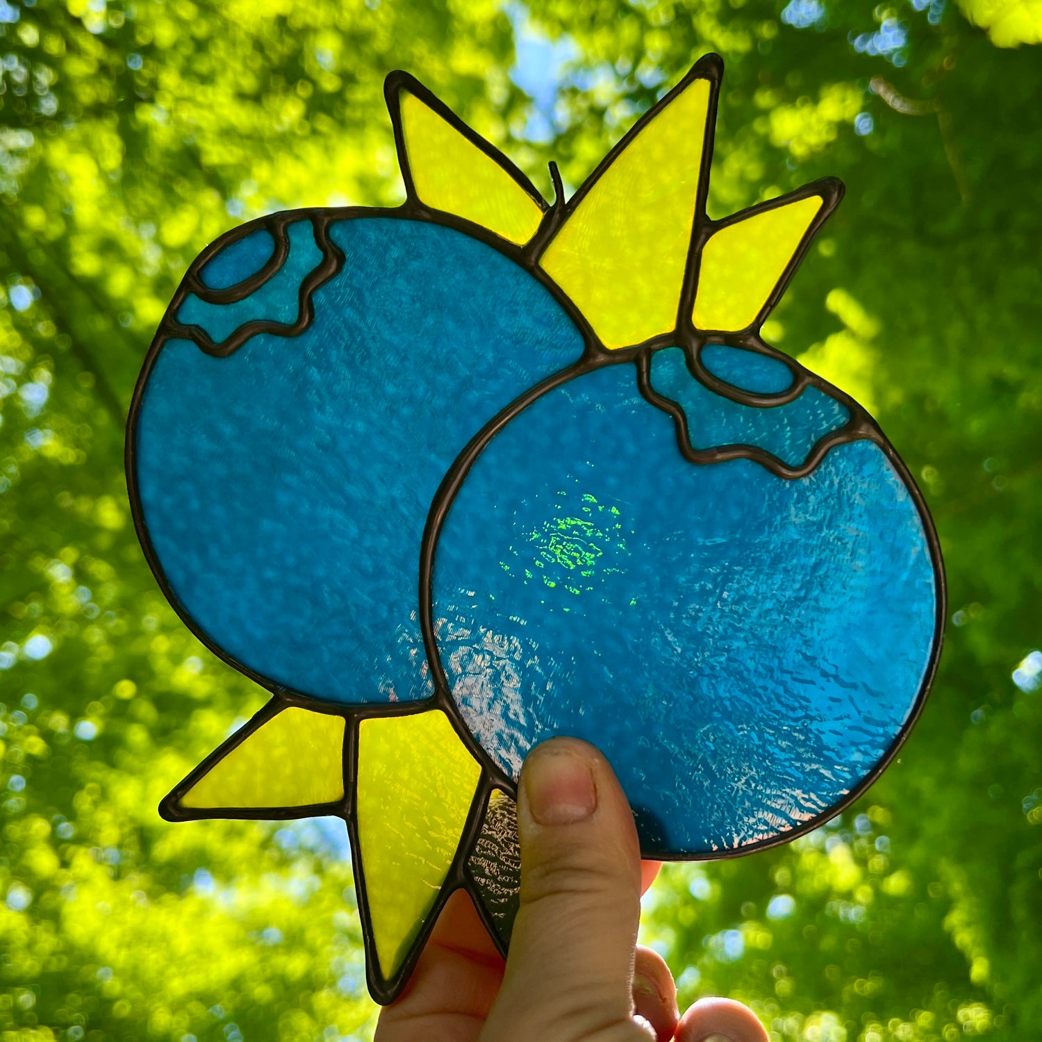 hand holding a dark blue stained glass suncatcher depicting a small bunch of blueberries with leaves in front of bright sunlight with bright green leaves and trees in the background