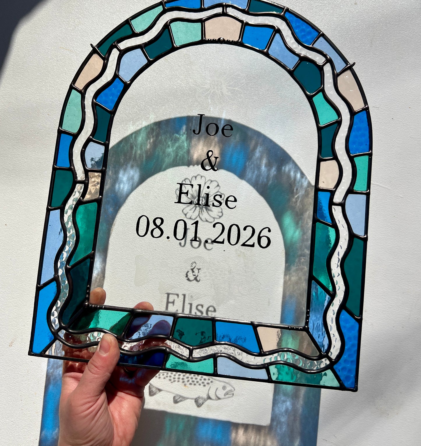 Stained glass panel in the shape of an arch with a multicolor blue, teal, turquoise, and pink border. Within the border is squiggle detail in clear textured glass. The center of the arch is clear uncolored glass, with personalized text and hand etched details of a cosmos flower and trout. The piece is held by a hand against a white wall, with sunlight casting refractions and shadows of the text, colored glass, and etchings.
