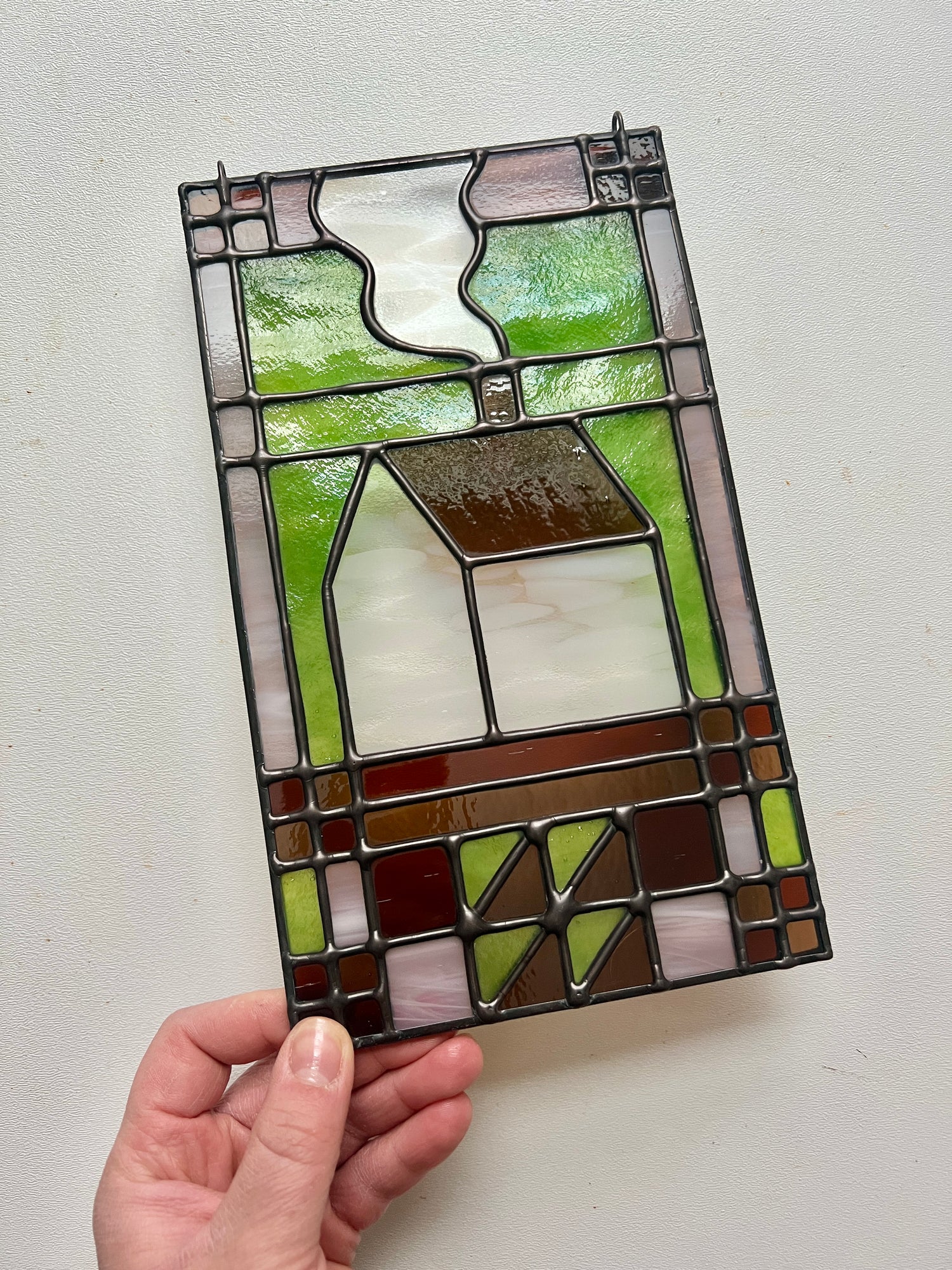 Stained glass textile inspired panel depicting a home in the center in shades of green, pink, white, and brown, held by a hand against a white background