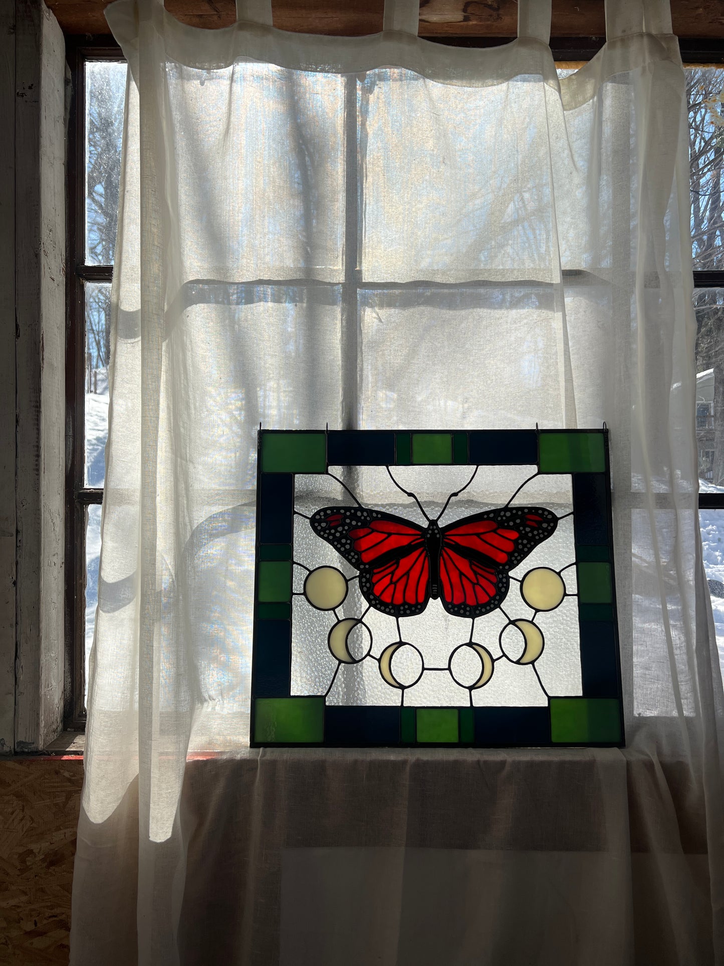 A large stained glass panel with a thick blue and green geometric border and a monarch butterfly and moon phases in the center, with textured clear glass in the background. The panel is resting against a window with a sheer curtain just behind it, offering gentle backlighting.