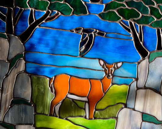 close up of a large stained glass panel depicting mossy, rocky cliffs and trees, with a deer, a kingfisher, and a raven standing amongst a river, greenery, and bright blue skies with some clouds and a bright yellow orange sun.